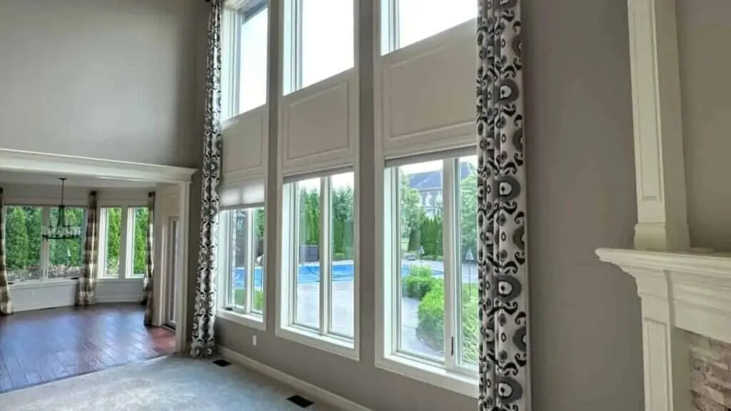 Two-story great room with warm gray walls, white window trim and a white fireplace mantel beside tall windows